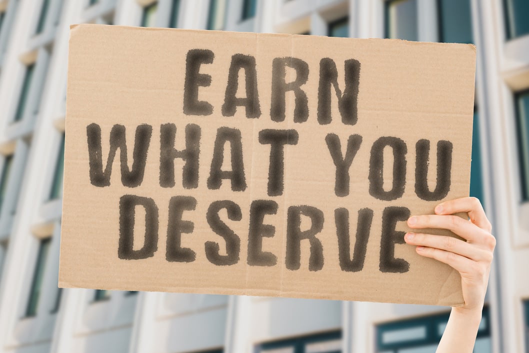 The phrase " Earn What You Deserve " on a banner in men's hand with blurred background. Win. Real. Salary. Wage. Price. Profit. Job. Step. Effort. Trophy. Do. More. Work. Self. Career. Victory. Target